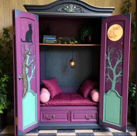 Decorative wardrobe with open doors showing a cozy interior with cushions and a small table. Decorative cabinet with a purple interior featuring a cat, moon, and books. Decorative wooden cabinet with open doors, featuring artistic designs and cushions. Upcycled Antique Witchy Cat Moon Armoire Reading Nook \| La Vie Nouveau Décor Shoppe