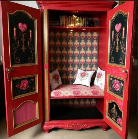 Decorative red cabinet with open doors, cushioned seat, and floral patterns. Decorative red cabinet with floral wallpaper, cushions, and books inside. Red ornate wardrobe with floral designs converted into a sitting area with cushions and pillows. Upcycled Antique Romantic Hearts Armoire Reading Nook \| La Vie Nouveau Décor Shoppe
